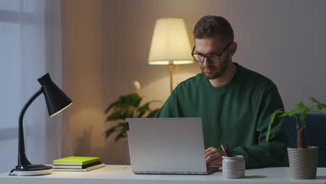 Der-Junge-Student-Lernt-Abends-Zu-Hause-Online,-Trägt-Eine-Brille,-Schaut-Auf-Den-Bildschirm-Und-Liest-Informationen-Und-Tippt-Nachrichten